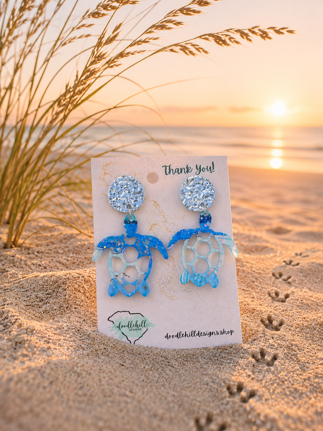 "Watercolor sea turtle acrylic earrings with silver glitter accents displayed on beach sand with turtle tracks and sea oats at sunrise."